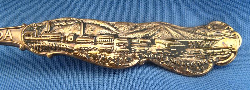 Souvenir Mining Spoon Copper Flat Handle Marking.JPG - SOUVENIR MINING SPOON COPPER FLAT ELY NEVADA - Copper souvenir mining spoon, embossed scene of open pit copper mine in bowl with marking COPPER FLAT at bottom, handle is a skyline depiction of mountains with buildings and smokestacks and a bridge with ELY NEVADA marked on handle, Steptoe Mining and Smelting Co. is marked at the bottom of the skyline, reverse has Paye & Baker makers mark with Sterling x’ed out, 5 1/4 in. long, weight 21 grams [Copper Flat is today a general reference to the area in eastern Nevada where the great copper discoveries were made at the first of the 20th century.  Copper Flat was also small settlement for a short time in this area (now long gone) approximately 6 miles west of Ely, NV in White Pine County.  The Copper Flat area is also associated with several small towns including Kimberly, Riepetown, Ruth and Veteran. The famed open-pit copper mines of eastern Nevada, including the Liberty Pit (largest in the state), are located in this area between Ely and Ruth just south of Highway 50. Through the first half of the 20th century, this area produced nearly a billion dollars in copper, gold and silver. The first mining claims were filed in White Pine County, Nevada, as early as 1867, and the first copper claim was filed in the summer of 1900.  Over the next couple years, an additional 26 claims covering 437 acres of the Copper Flat area were filed.  The Nevada Consolidated Copper Company was organized and incorporated under the laws of Maine on November 17, 1904 to buy up and operate these claims. By May 1906, the company was controlled by the Guggenheim family and their associates and in 1907, steam shovels began stripping the overburden above the Eureka mine to start the open pit mining operations.  In 1906, the Nevada Consolidated Copper Company and the Cumberland and Ely Mining Corporation formed a partnership to build a smelter in the area to process the ores from Copper Flat.  Named the Steptoe Valley Mining and Smelting Company, the smelter started construction in December 1906 at the small town of McGill some 12 miles north of Ely.  The smelter was completed and operations began on May 15, 1908.  The first copper was shipped from the smelter on August 7 of the same year. Kennecott Utah Copper acquired Nevada Consolidated Copper Company, which included Steptoe Valley Mining and Smelting Company and the smelter at McGill, in 1932. In 1983, the price of copper along with the low grade ore being mined led to Kennecott closing the smelter and demolishing it.]